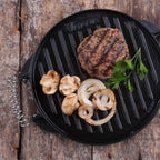 Vista superior de la plancha redonda reversible Victoria de 32 cm en hierro fundido curado, mostrando su lado tipo parrilla con una hamburguesa, champiñones y cebollas asadas sobre una superficie de madera - Almacén La Palma - 1050070