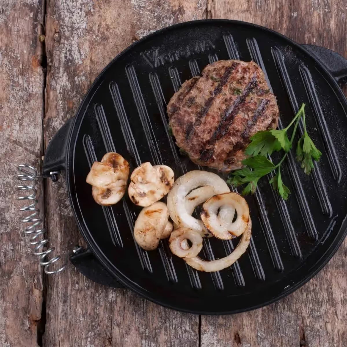 Vista superior de la plancha redonda reversible Victoria de 32 cm en hierro fundido curado, mostrando su lado tipo parrilla con una hamburguesa, champiñones y cebollas asadas sobre una superficie de madera - Almacén La Palma - 1050070
