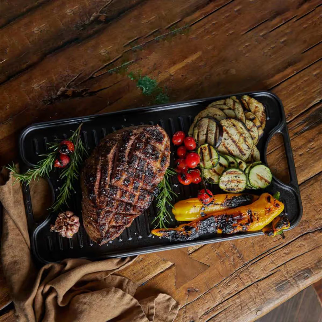 Vista cenital de la cara estriada tipo grill de la plancha reversible Victoria de 47x25 cm en hierro fundido curado con carne asada, vegetales a la parrilla, romero y ajo sobre una mesa de madera rústica - Ferretería La Palma - 1050023