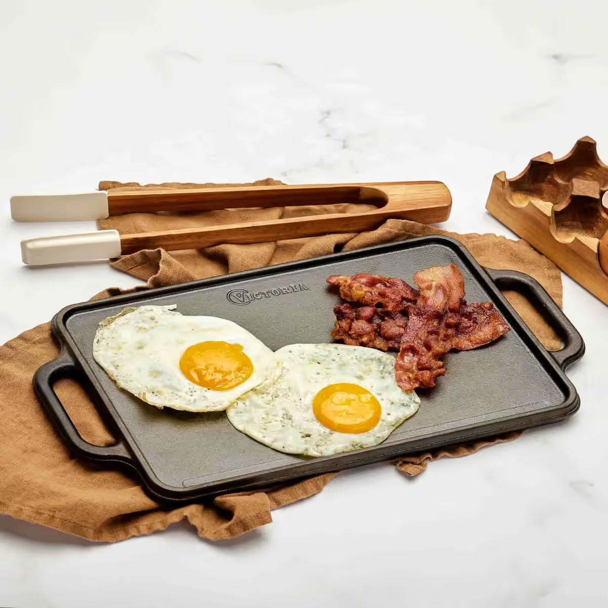 Vista en perspectiva de la cara lisa de la plancha reversible Victoria de 33x21 cm en hierro fundido curado con huevos fritos y tocino servidos sobre un paño café en una mesa blanca - Ferretería La Palma - 1050091
