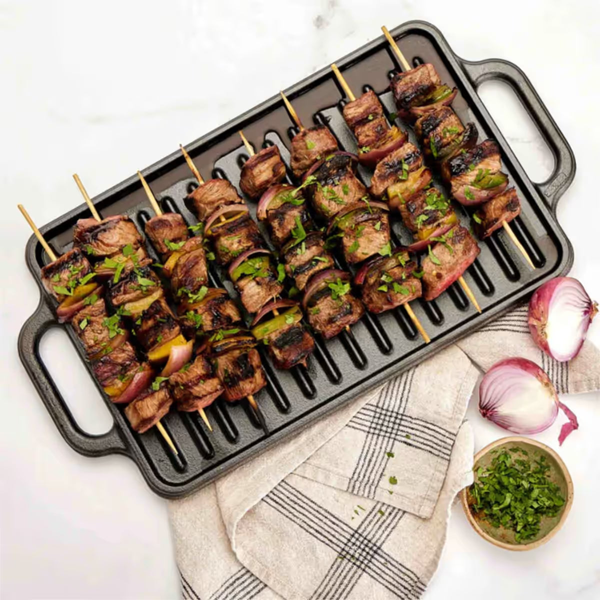 Vista cenital de la cara estriada tipo grill de la plancha reversible Victoria de 33x21 cm en hierro fundido esmaltado con brochetas de carne y vegetales sobre una mesa blanca - Ferretería La Palma - 1050020