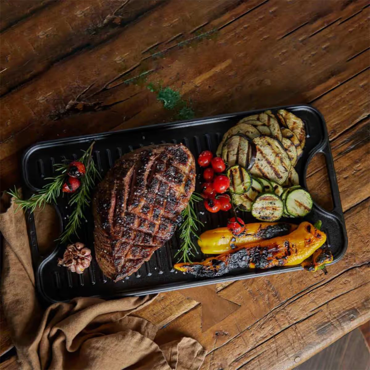 Vista cenital de la cara estriada tipo grill de la plancha reversible Victoria de 47x25 cm en hierro fundido esmaltado con carne asada, vegetales a la parrilla, romero y ajo sobre una mesa de madera rústica - Ferretería La Palma - 1050023
