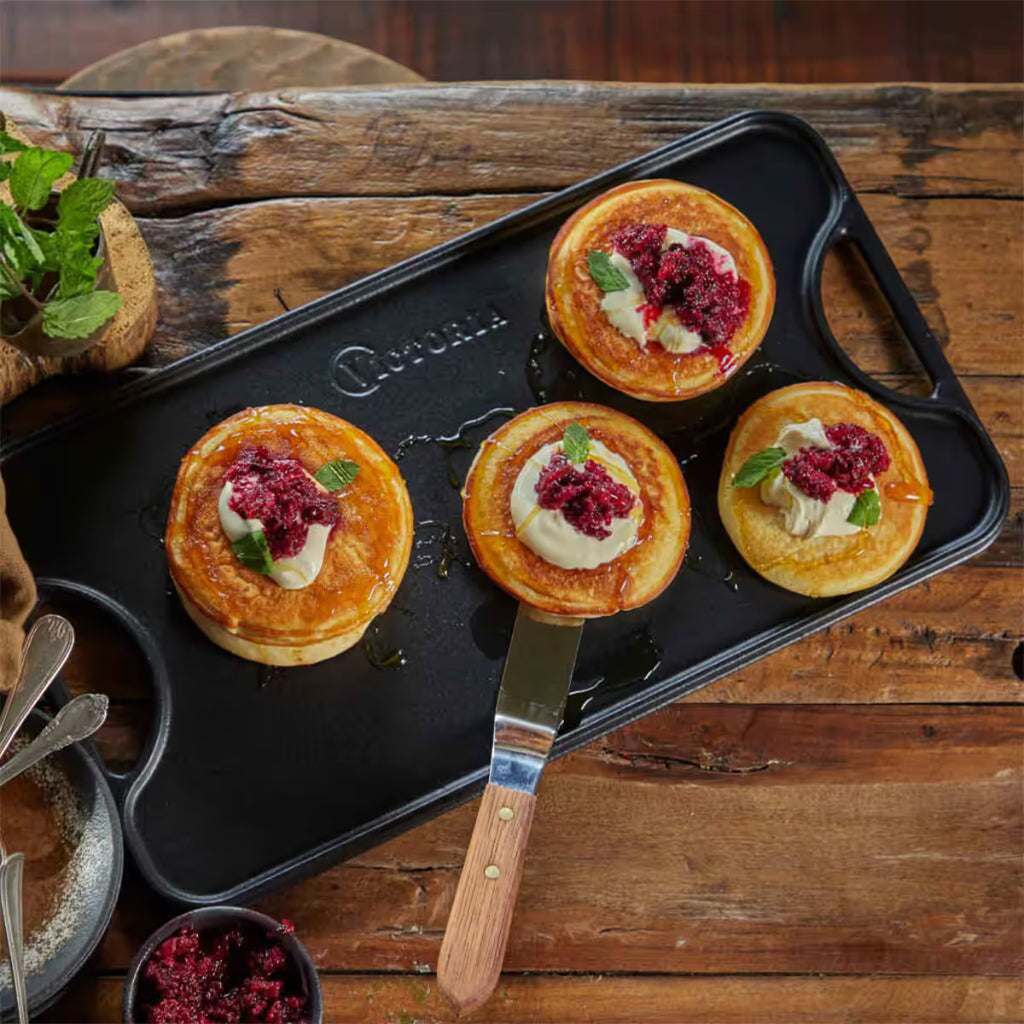 Vista en perspectiva de la cara lisa de la plancha reversible Victoria de 47x25 cm en hierro fundido esmaltado con pancakes con crema, frutos rojos y miel servidos sobre una mesa de madera rústica - Ferretería La Palma - 1050023