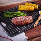 Vista en perspectiva de la cara estriada tipo grill de la plancha reversible Victoria de 50x35 cm en hierro fundido esmaltado con carne asada, espárragos y mazorcas de maíz sobre una mesa de madera - Ferretería La Palma - 1050082