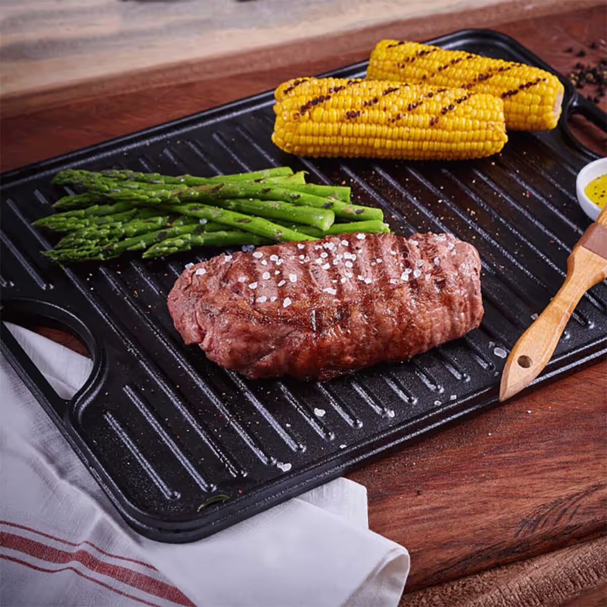 Vista en perspectiva de la cara estriada tipo grill de la plancha reversible Victoria de 50x35 cm en hierro fundido esmaltado con carne asada, espárragos y mazorcas de maíz sobre una mesa de madera - Ferretería La Palma - 1050082