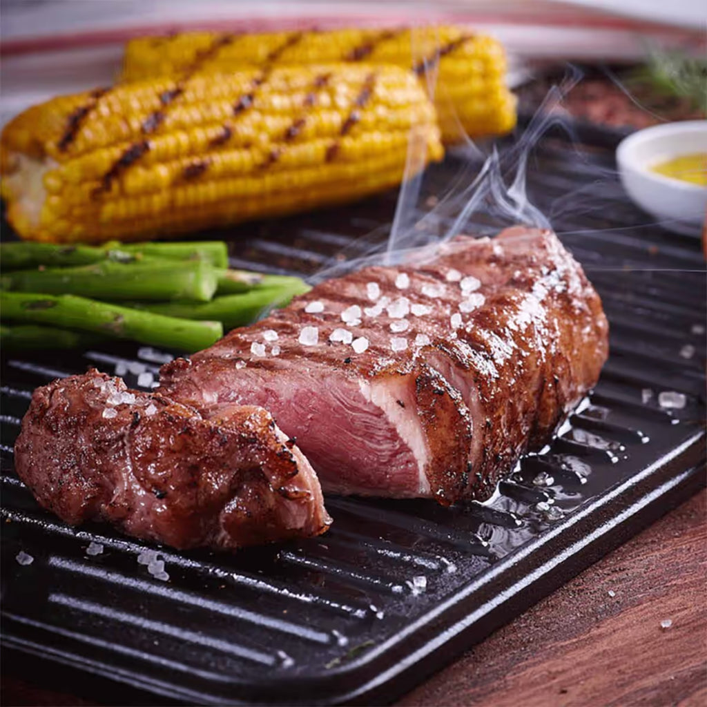 Vista de primer plano de la cara estriada tipo grill de la plancha reversible Victoria de 50x35 cm en hierro fundido esmaltado con un corte de carne asada humeante, espárragos y mazorcas de maíz sobre una mesa de madera - Ferretería La Palma - 1050082