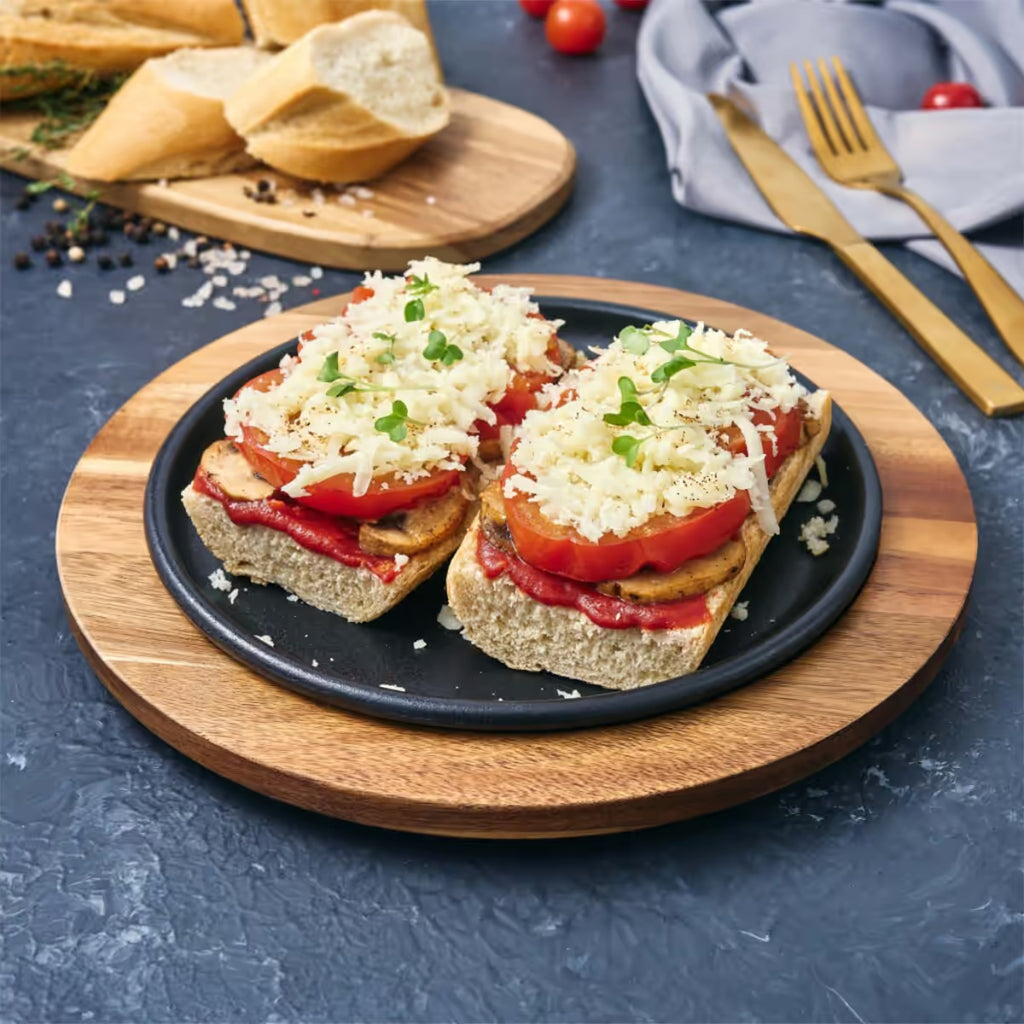 Vista en perspectiva del plato redondo Victoria de 20 cm en hierro fundido esmaltado con panes con tomate, champiñones y queso servidos sobre una base de madera en una mesa gris - Ferretería La Palma - 1050036