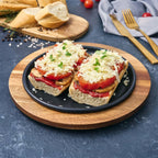 Vista en perspectiva del plato redondo Victoria de 20 cm en hierro fundido esmaltado con panes con tomate, champiñones y queso servidos sobre una base de madera en una mesa gris - Ferretería La Palma - 1050036