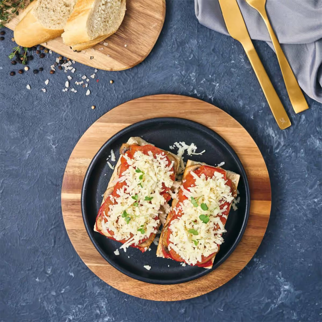 Vista cenital del plato redondo Victoria de 20 cm en hierro fundido esmaltado con panes con tomate, champiñones y queso servidos sobre una base de madera en una mesa gris - Ferretería La Palma - 1050036