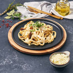 Vista en perspectiva del plato redondo Victoria de 26 cm en hierro fundido esmaltado con pasta y pollo servidos sobre una base de madera en una mesa gris - Ferretería La Palma - 1050037