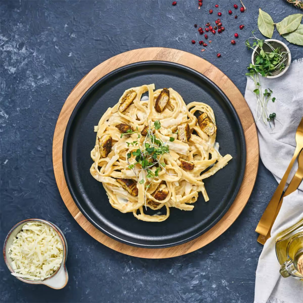 Vista cenital del plato redondo Victoria de 26 cm en hierro fundido esmaltado con pasta y pollo servidos sobre una base de madera en una mesa gris - Ferretería La Palma - 1050037
