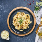 Vista cenital del plato redondo Victoria de 26 cm en hierro fundido esmaltado con pasta y pollo servidos sobre una base de madera en una mesa gris - Ferretería La Palma - 1050037