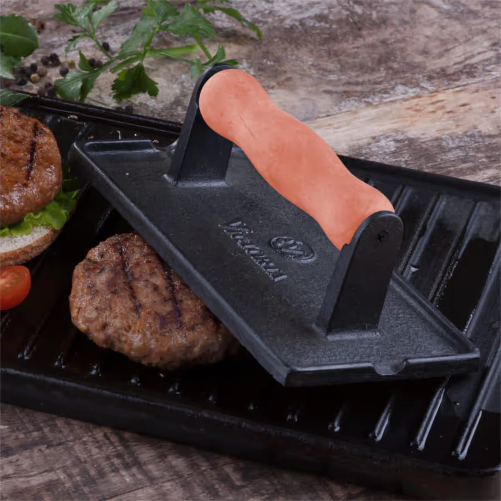 Vista superior en perspectiva de la prensa rectangular Victoria de 21 cm en hierro fundido curado con mango de madera siendo utilizada para sellar una hamburguesa sobre una plancha tipo grill - Ferretería La Palma - 1050065