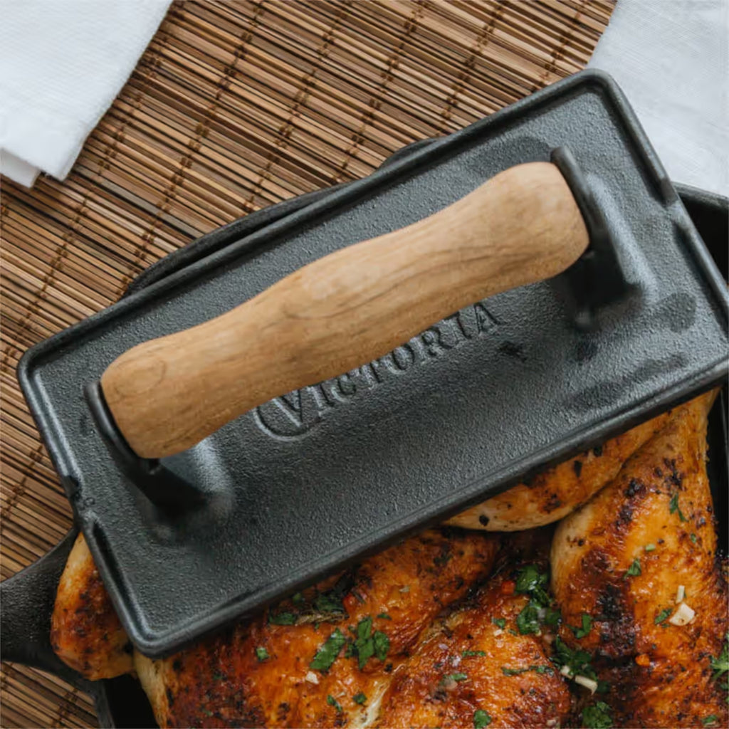Vista superior en perspectiva de la prensa rectangular Victoria de 21 cm en hierro fundido curado con mango de madera siendo utilizada para presionar un pollo asado sobre un individual de bambú - Ferretería La Palma - 1050065