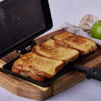 Vista de la sandwichera manual Victoria en hierro fundido esmaltado abierta con tres tajadas de pan tostado en su interior, ambientada sobre una base de madera junto a una manzana verde - Almacén La Palma - 1050105