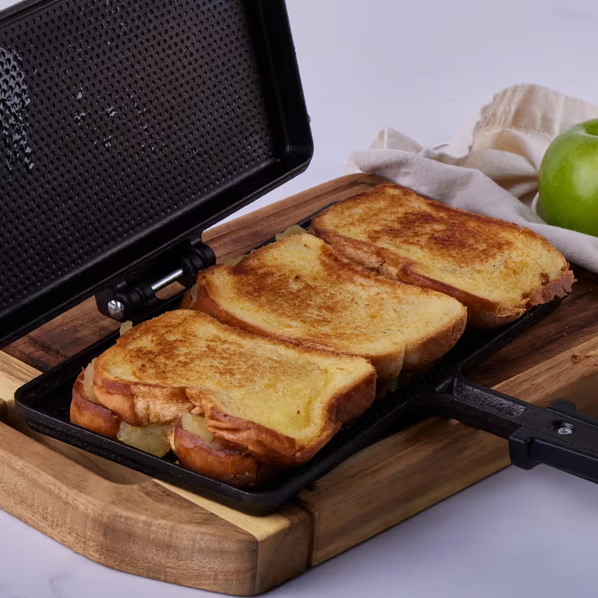 Vista de la sandwichera manual Victoria en hierro fundido esmaltado abierta con tres tajadas de pan tostado en su interior, ambientada sobre una base de madera junto a una manzana verde - Almacén La Palma - 1050105