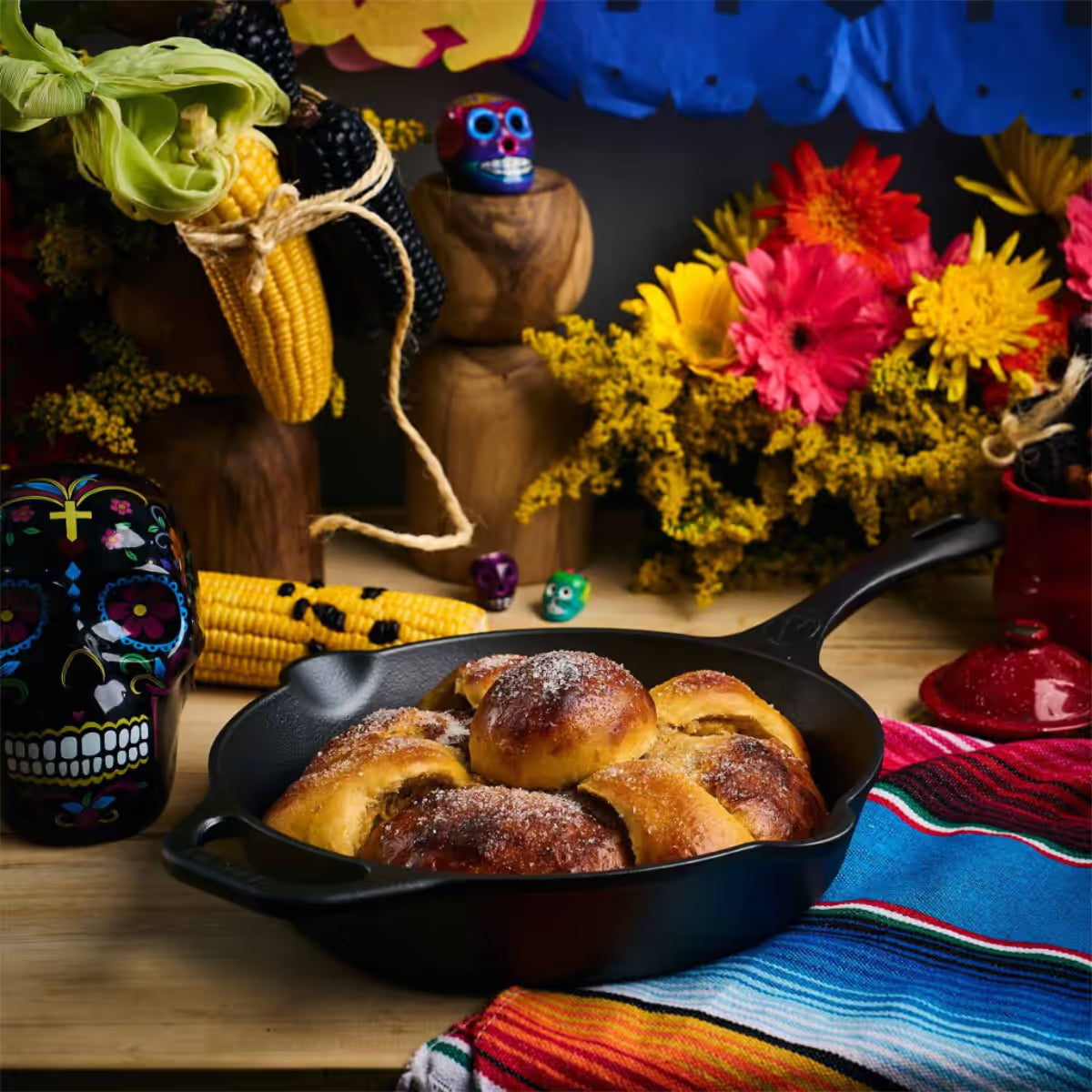 Vista en perspectiva del sartén de hierro fundido Victoria de 25 cm ambientado con pan de muerto, calaveras decorativas, mazorcas de maíz y flores sobre un mantel colorido - Almacén La Palma - 1050097