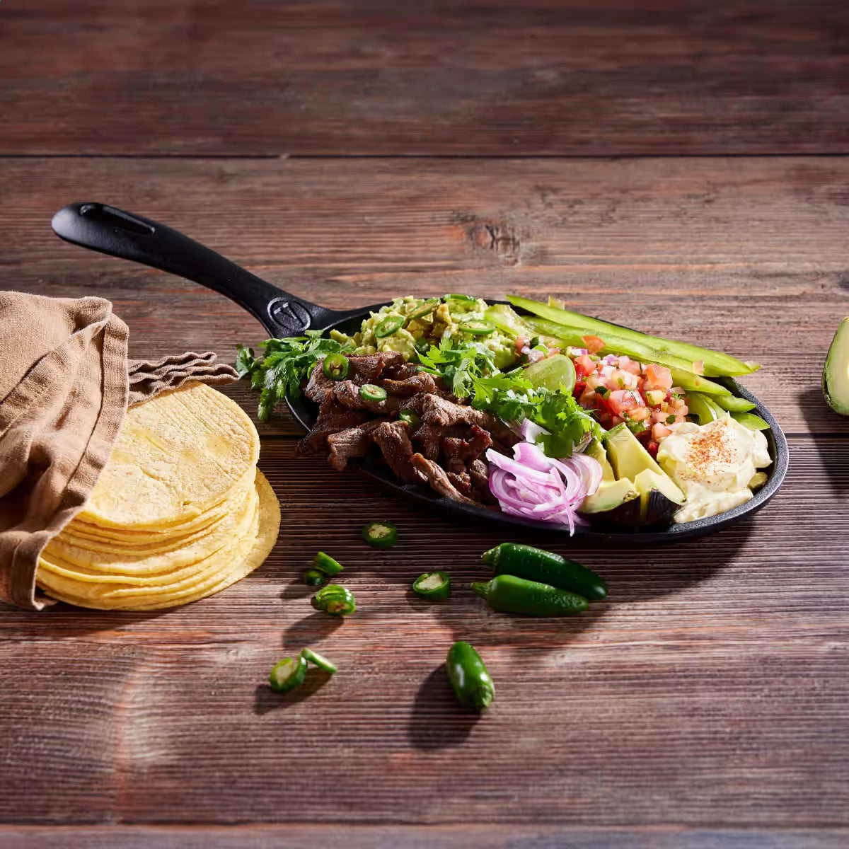 Sartén para fajitas Victoria esmaltado de 24 cm con carne asada, guacamole, vegetales y stack de tortillas sobre una mesa de madera rústica - Ferretería La Palma - 1050050