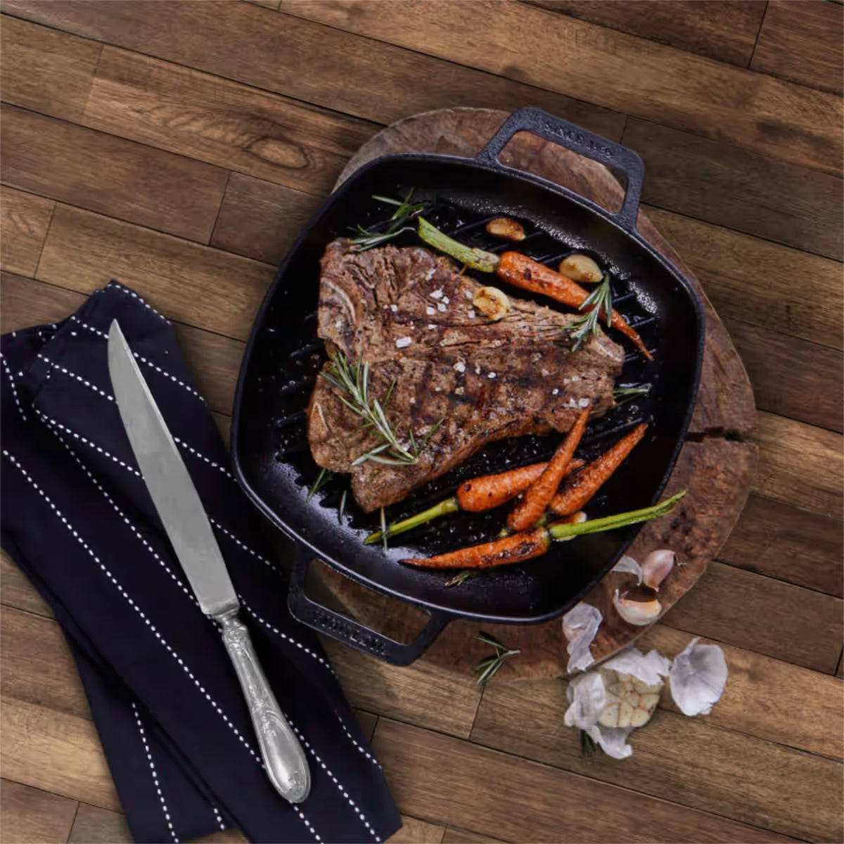Vista cenital del sartén grill cuadrado Victoria de 25 cm en hierro fundido curado con doble asa lateral con un corte de carne T-bone, zanahorias baby, ajos y romero fresco - Ferretería La Palma - 1050069