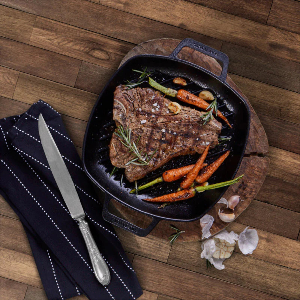 Vista cenital del sartén grill cuadrado Victoria de 25 cm en hierro fundido esmaltado con doble asa lateral con un corte de carne T-bone, zanahorias baby, ajos y romero fresco - Ferretería La Palma - 1050090