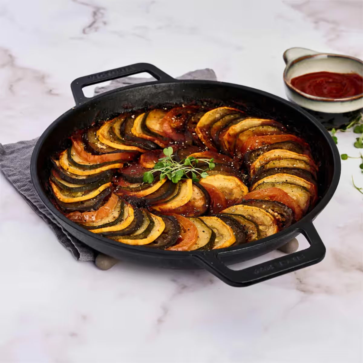 Sartén redondo Victoria de 25 cm en hierro fundido curado con doble asa lateral con un ratatouille de vegetales sobre una superficie de mármol - Ferretería La Palma - 1050068