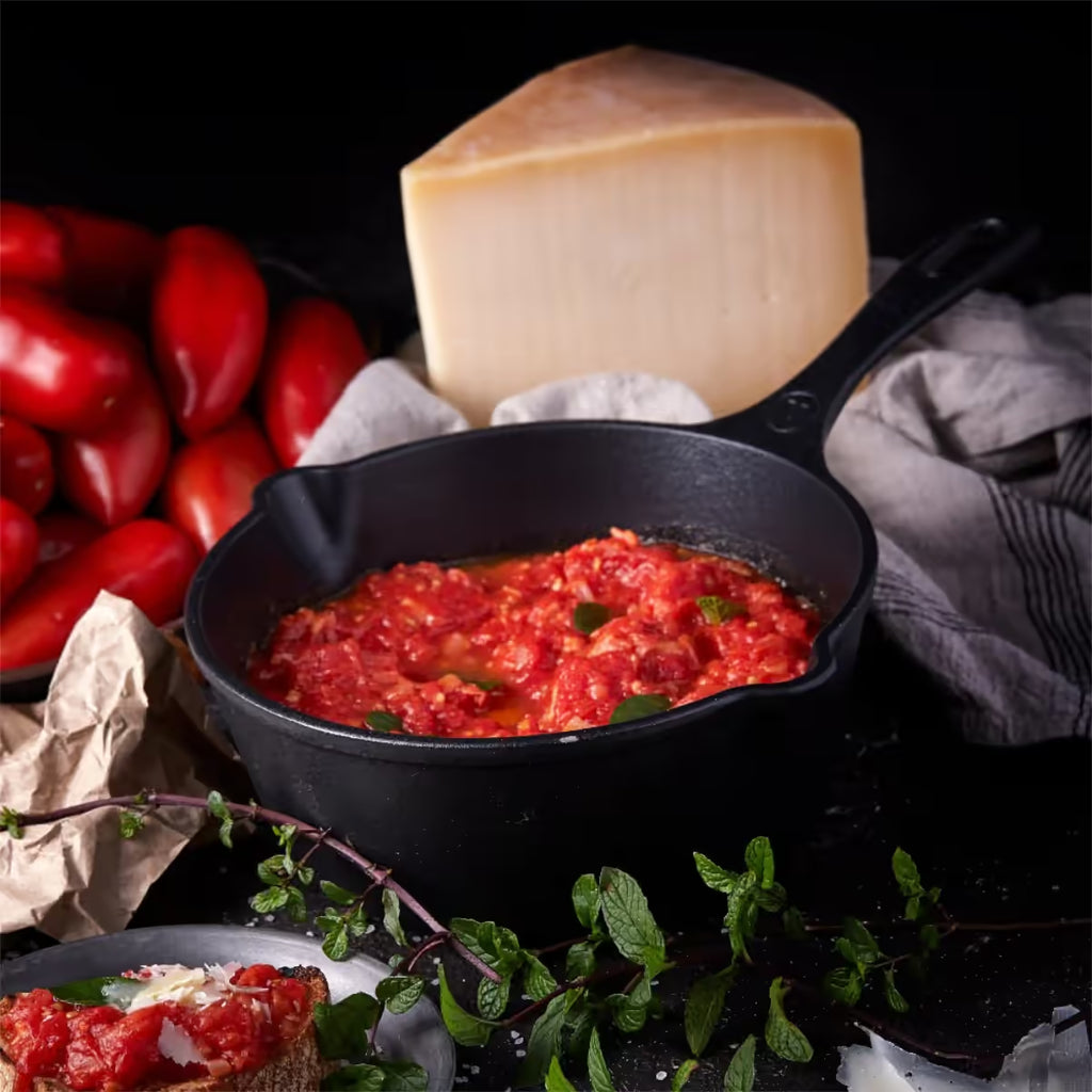 Vista en perspectiva del sartén profundo Victoria de 20 cm en hierro fundido esmaltado con capacidad de 1.9 litros con salsa de tomate, tomates frescos y queso sobre una mesa negra - Ferretería La Palma - 1050088