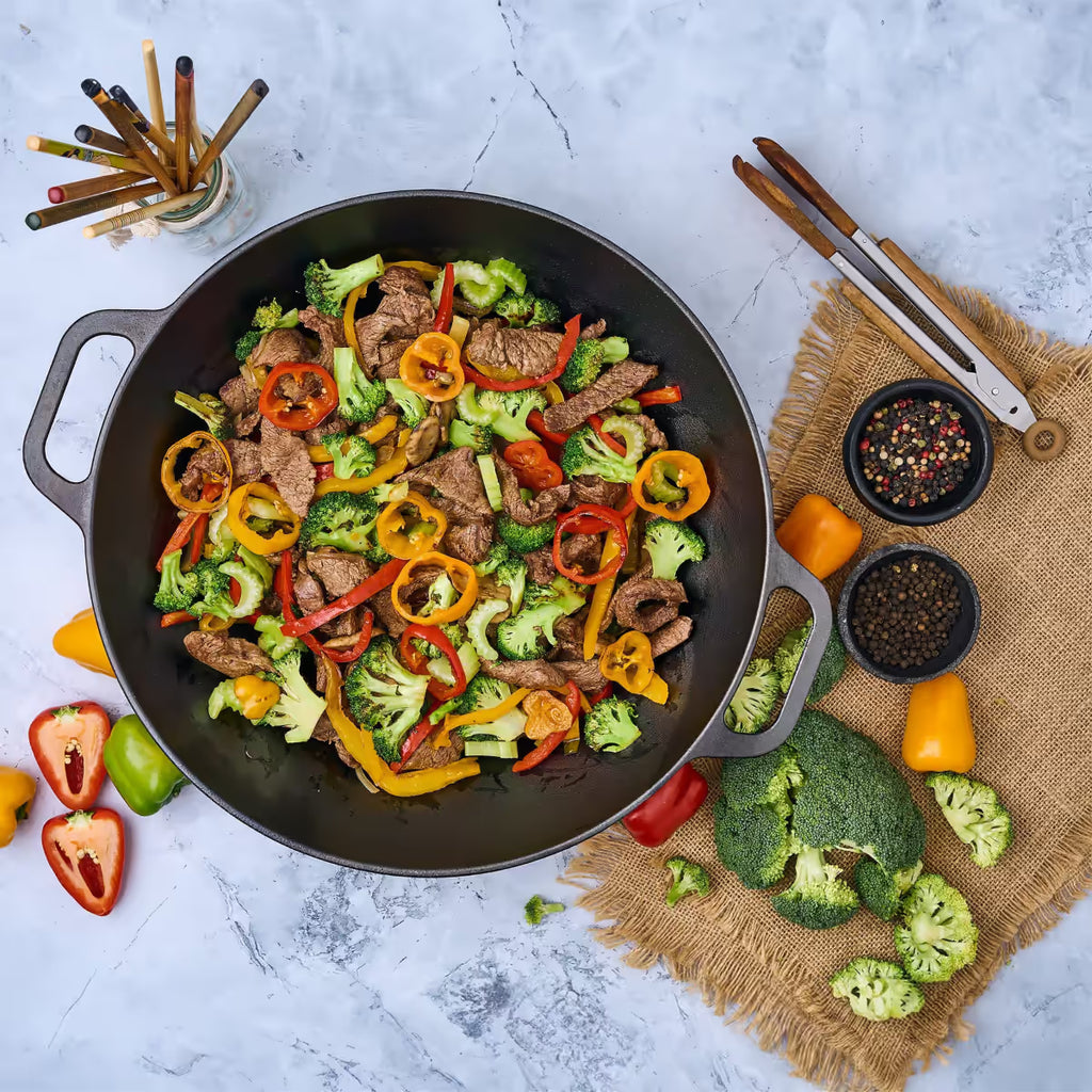 Vista cenital del sartén wok Victoria de 35.5 cm en hierro fundido curado con un salteado de carne, brócoli y pimientos rodeado de ingredientes frescos - Ferretería La Palma - 1050039