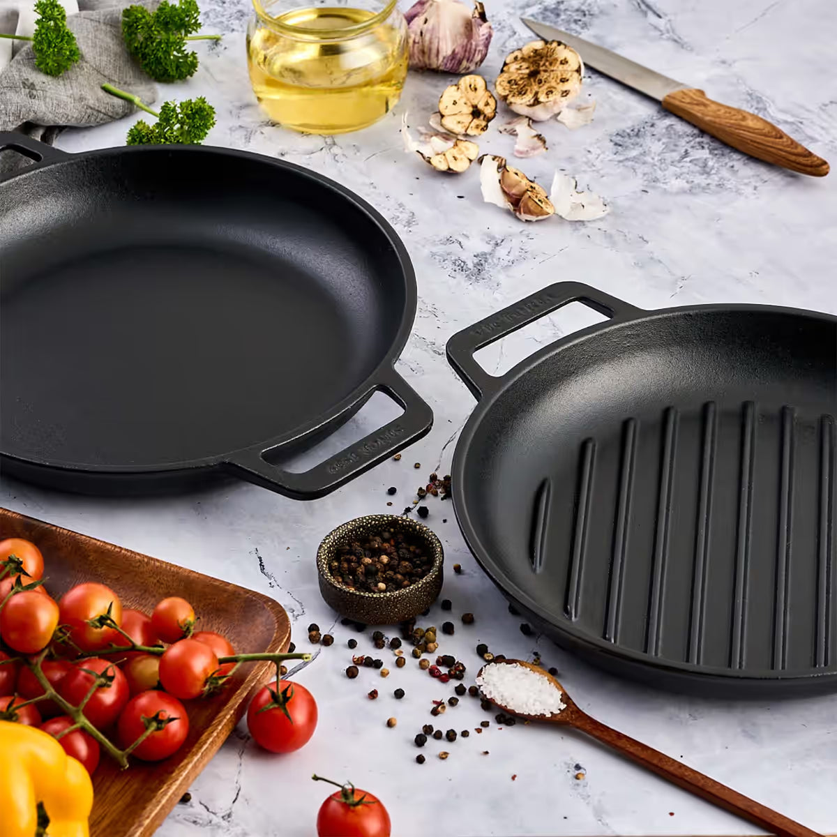 Vista en perspectiva del set de dos sartenes redondos Victoria de 25 cm en hierro fundido esmaltado con doble asa lateral, incluyendo un modelo liso y uno tipo grill, junto a ingredientes de cocina sobre una superficie de mármol - Ferretería La Palma - 1050149