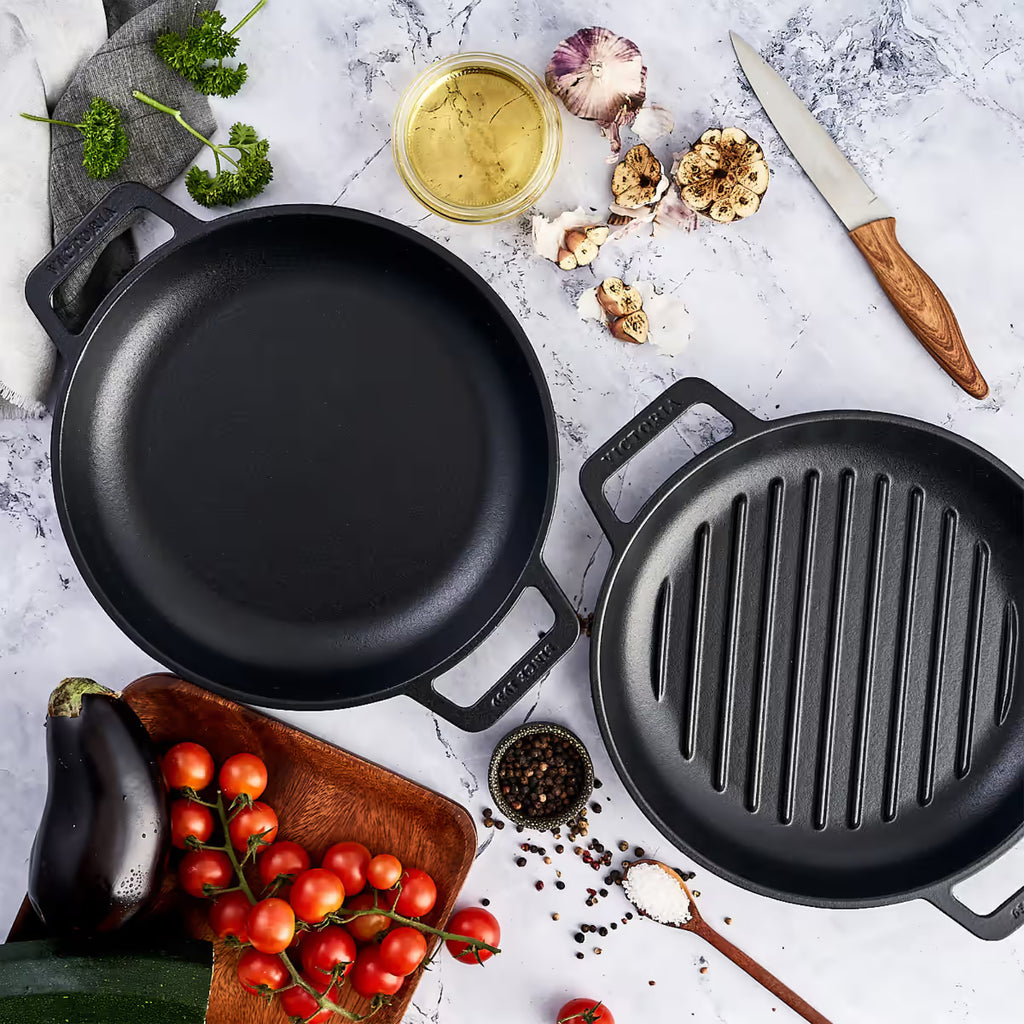 Vista cenital del set de dos sartenes redondos Victoria de 25 cm en hierro fundido esmaltado con doble asa lateral, incluyendo un modelo liso y uno tipo grill, rodeados de ingredientes de cocina sobre una superficie de mármol - Ferretería La Palma - 1050149