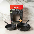 Vista del set de dos sartenes Victoria de 20 cm y 25 cm en hierro fundido esmaltado junto a su empaque original sobre una superficie de mármol - Ferretería La Palma - 1050099