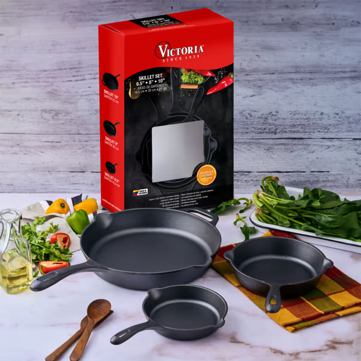 Vista del set de tres sartenes Victoria de 16.5 cm, 20 cm y 25 cm en hierro fundido esmaltado junto a su empaque original e ingredientes frescos sobre una superficie de mármol - Ferretería La Palma - 1050098
