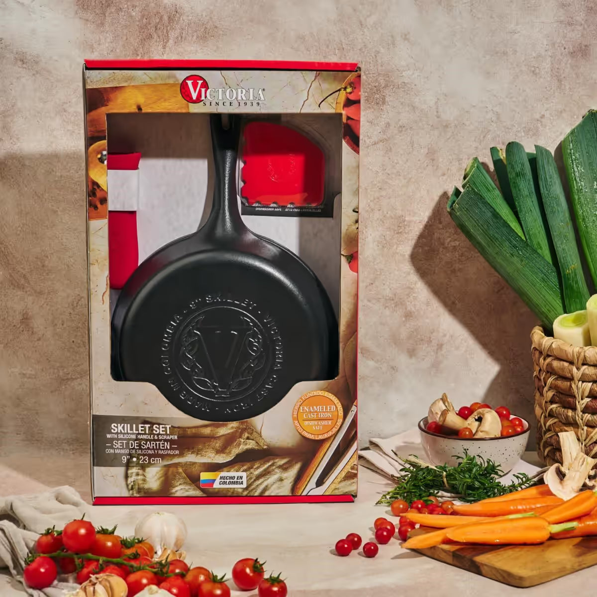 Vista frontal del empaque del set de sartén redondo Victoria de 23 cm en hierro fundido esmaltado que incluye protector de mango de silicona rojo y raspador junto a vegetales frescos - Ferretería La Palma - 1050148
