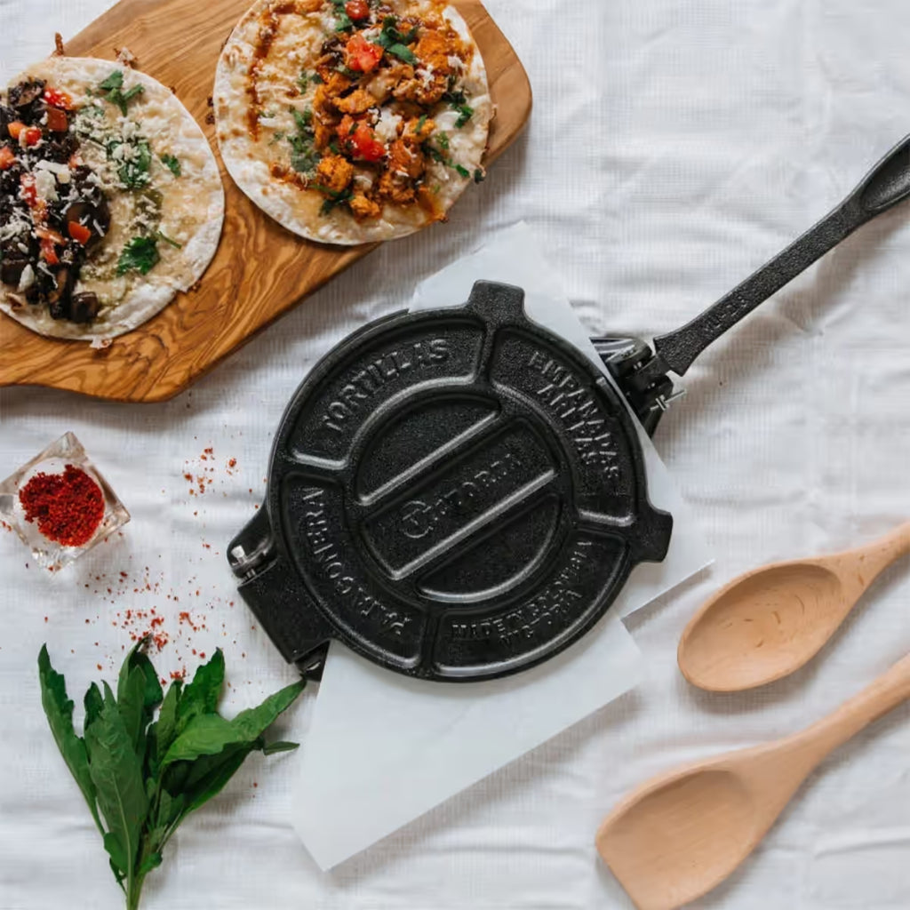 Vista cenital (superior) de la prensa para tortillas de hierro fundido marca Victoria en un ambiente de cocina con tacos preparados, hierbas frescas y utensilios de madera. Se aprecia el diseño circular de 16.5 centímetros (6.5 pulgadas) y el mango extendido de alta resistencia, ideal para preparaciones artesanales. Disponible en Ferretería La Palma - 1050010.